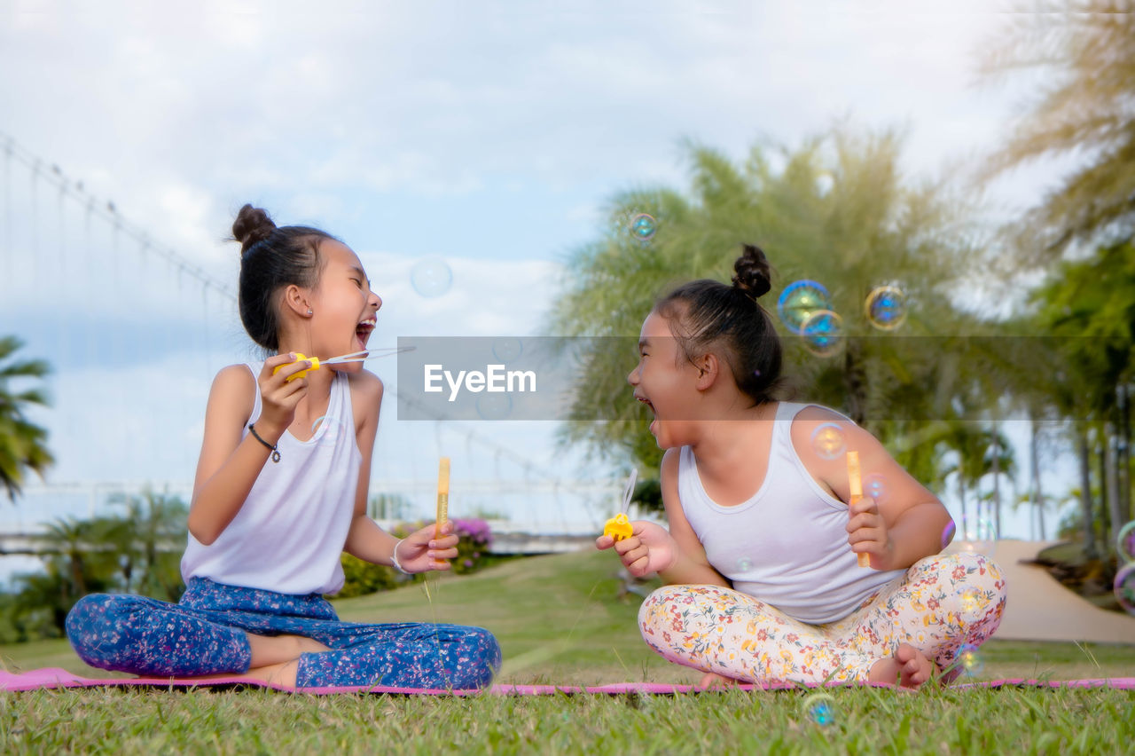 Cheerful girls playing with bubble wand while sitting on field against sky