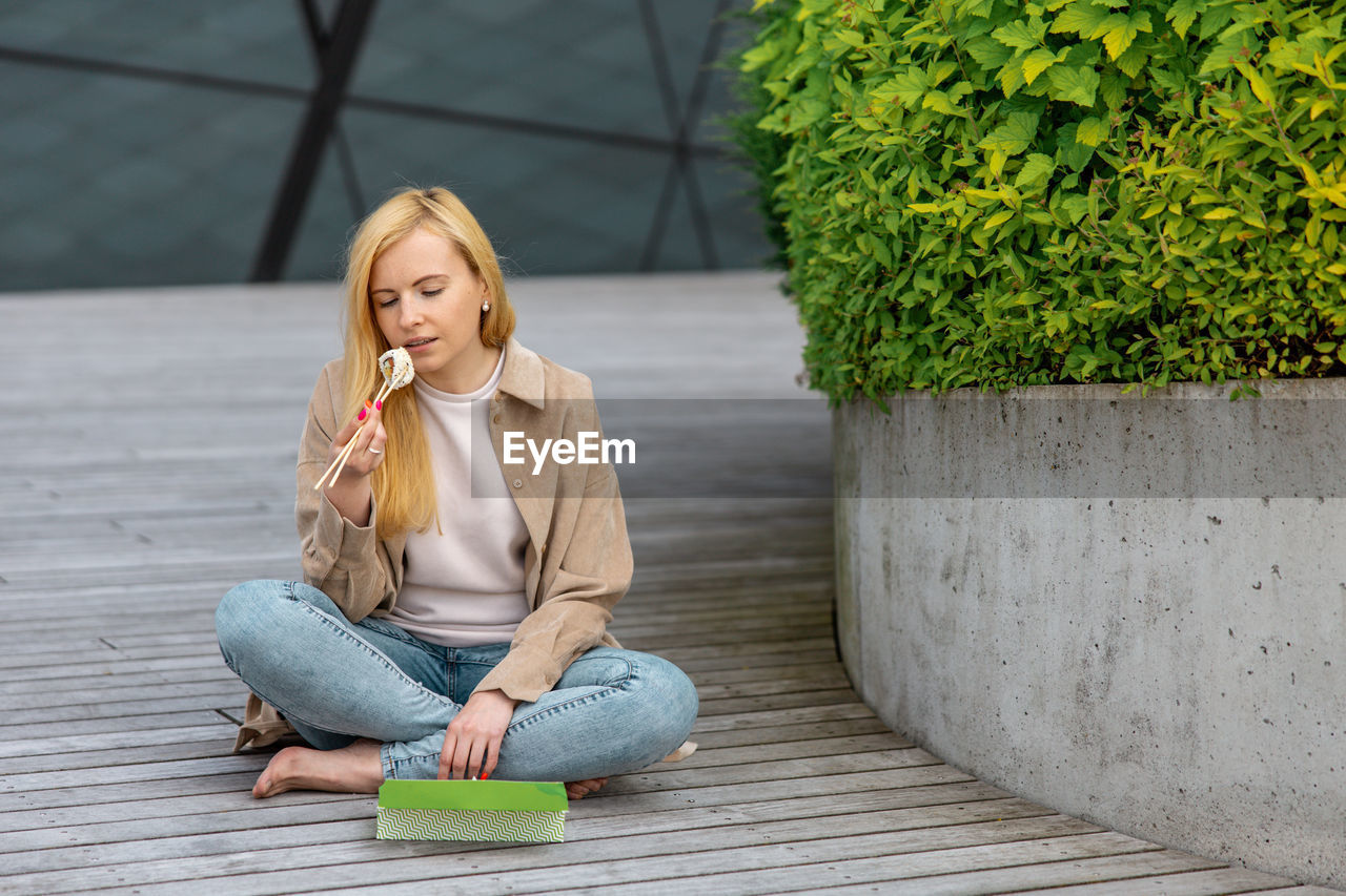 Young beautiful blond woman eating sushi outdoors, on the wooden terrace, by modern building