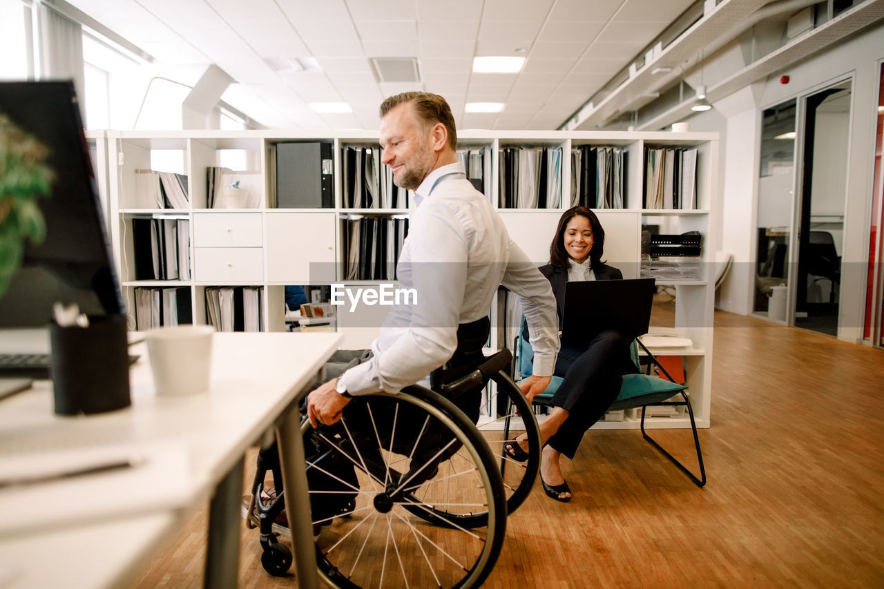 Disabled male professional sitting on wheelchair while smiling businesswoman using laptop at work place