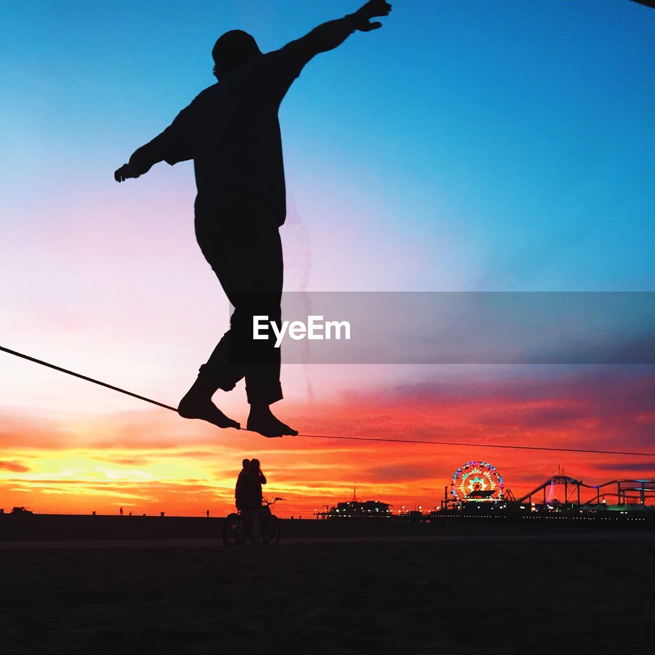 Low angle view of silhouette man walking on rope against cloudy sky during sunset