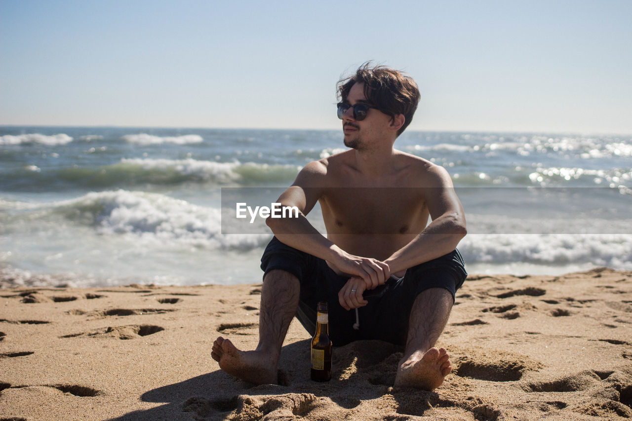 Shirtless young man sitting on beach against sky