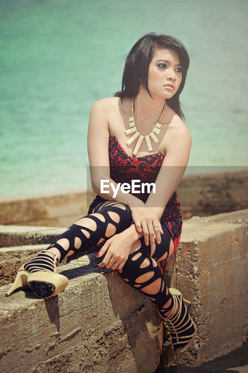 PORTRAIT OF BEAUTIFUL YOUNG WOMAN SITTING ON RETAINING WALL AGAINST SEA