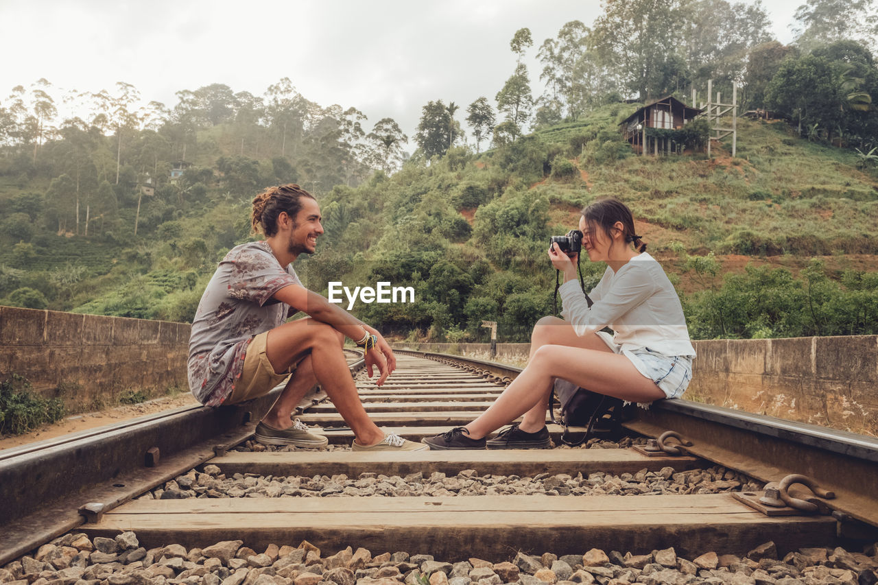Side view of young male and female travelers sitting on railroad in front of each other and taking photo on camera with green plants on background in sri lanka asian