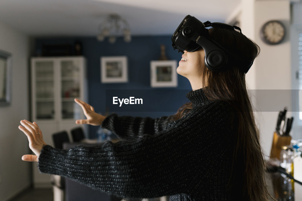 Playful teenage girl wearing virtual reality simulator eyeglasses sitting at home