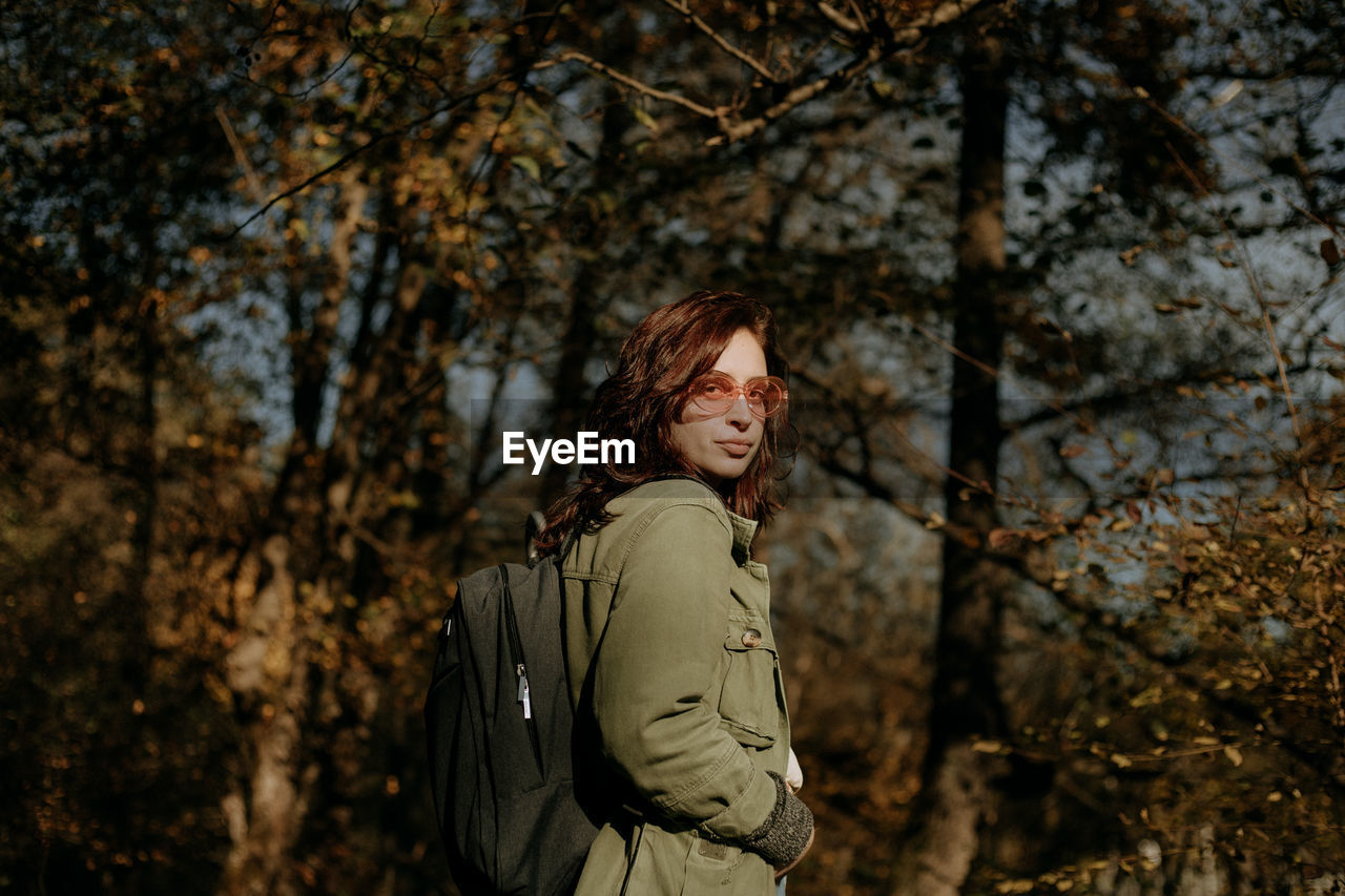 Young woman looking away while standing next to a tree in forest during autumn 