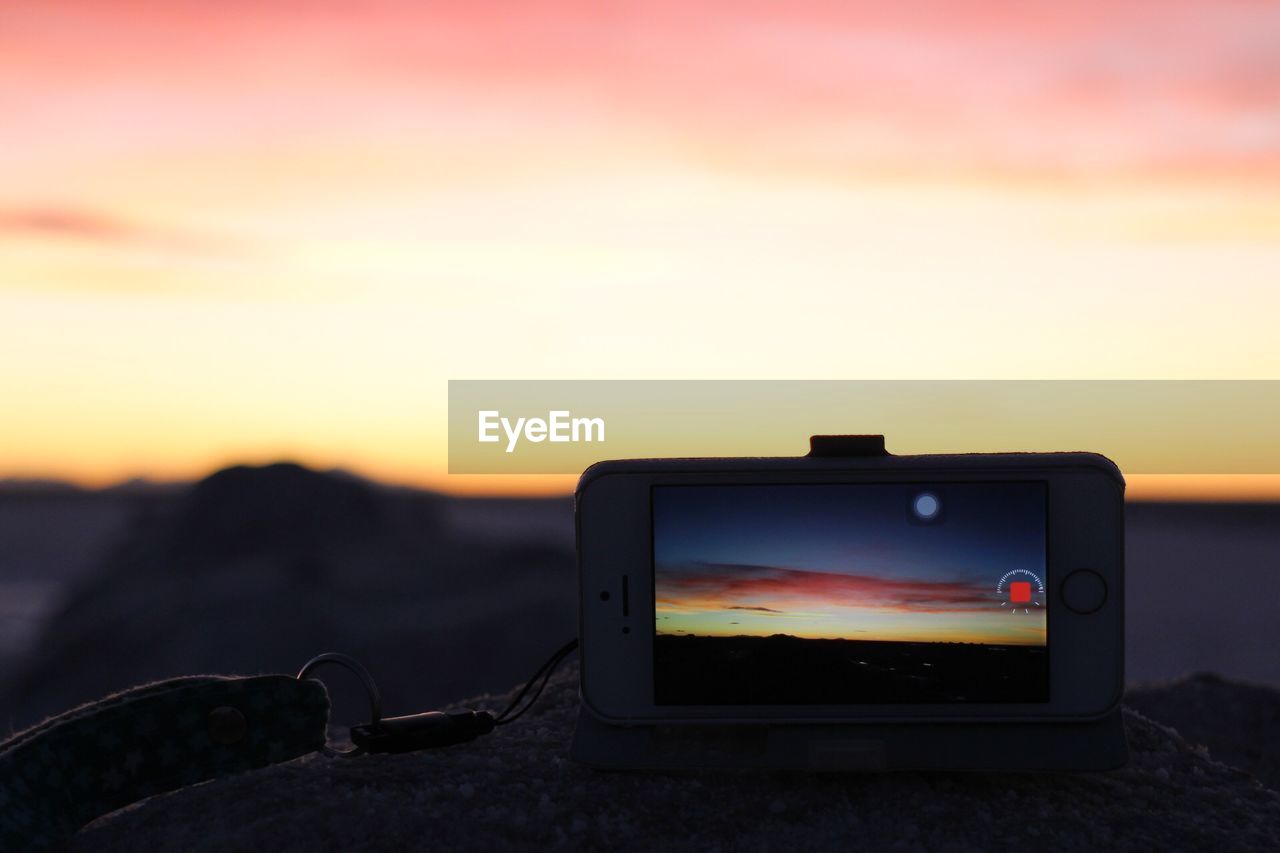 Close-up of smart phone on rock against sky during sunset