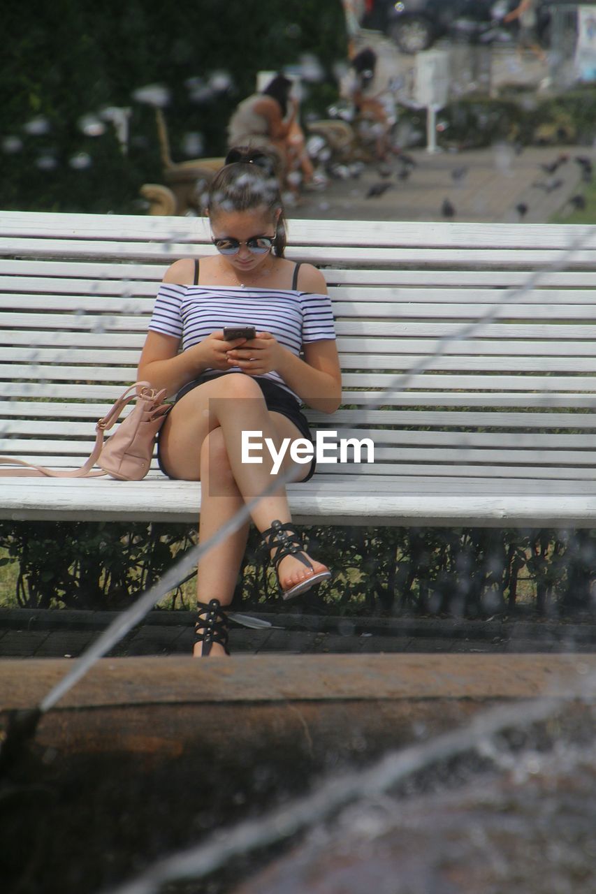 Full length of woman using phone while sitting on bench at park