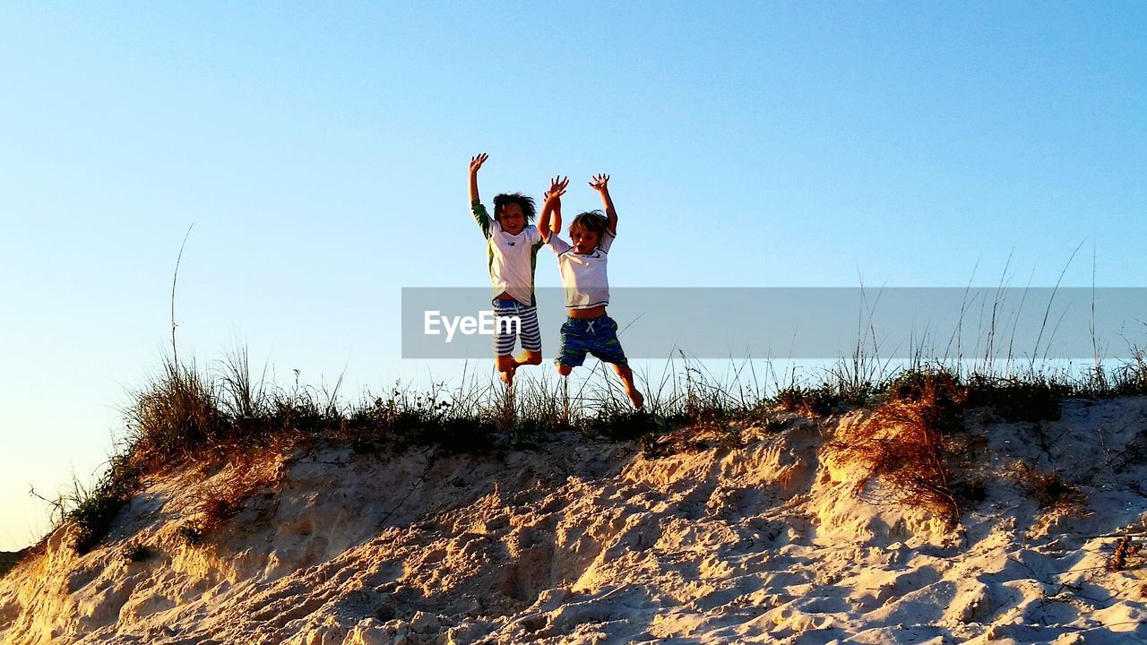 Low angle view of brothers jumping on sand hill at beach against clear sky