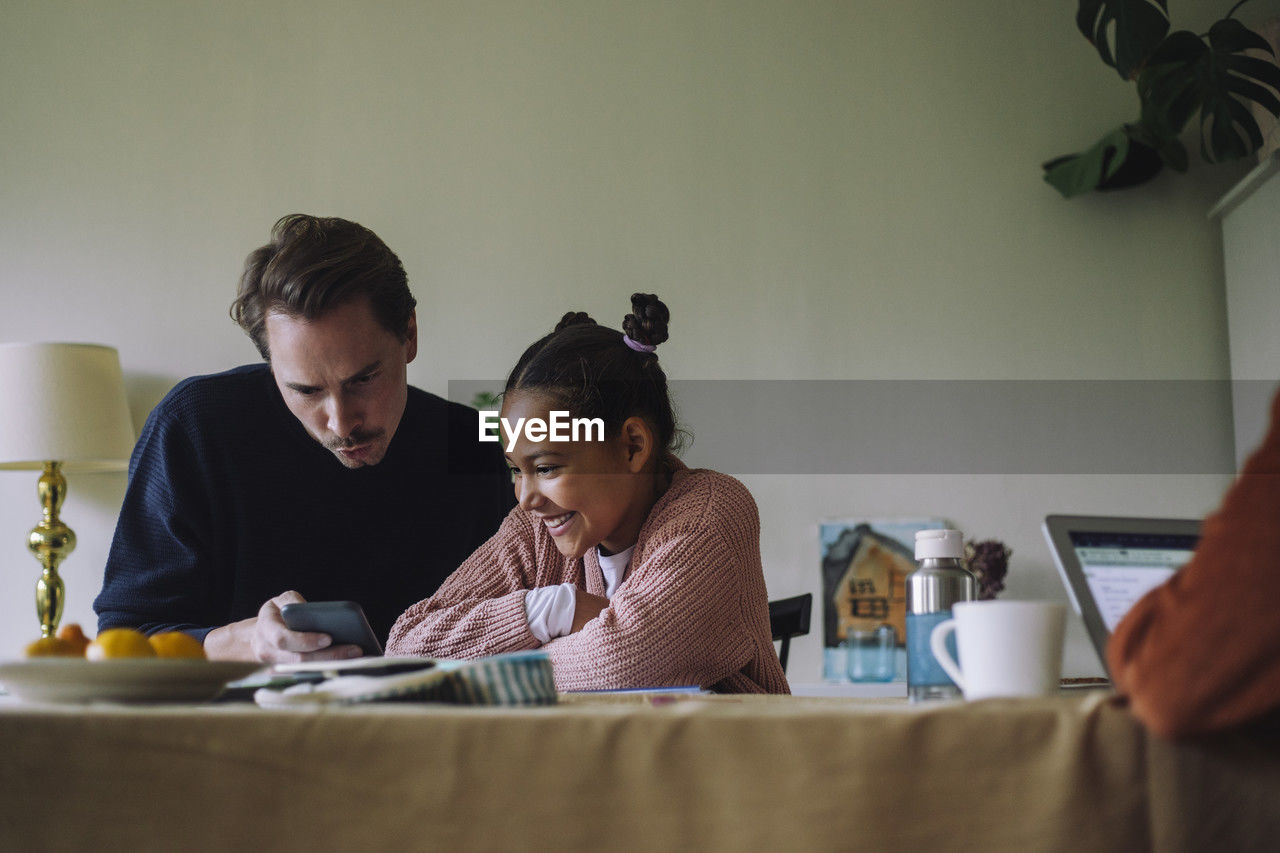 Gay man sharing smart phone with happy daughter sitting at dining table in home