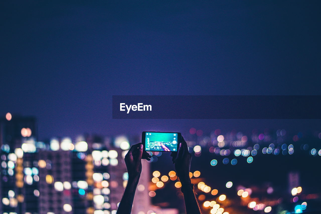 Cropped image of hand photographing illuminated cityscape against sky at night