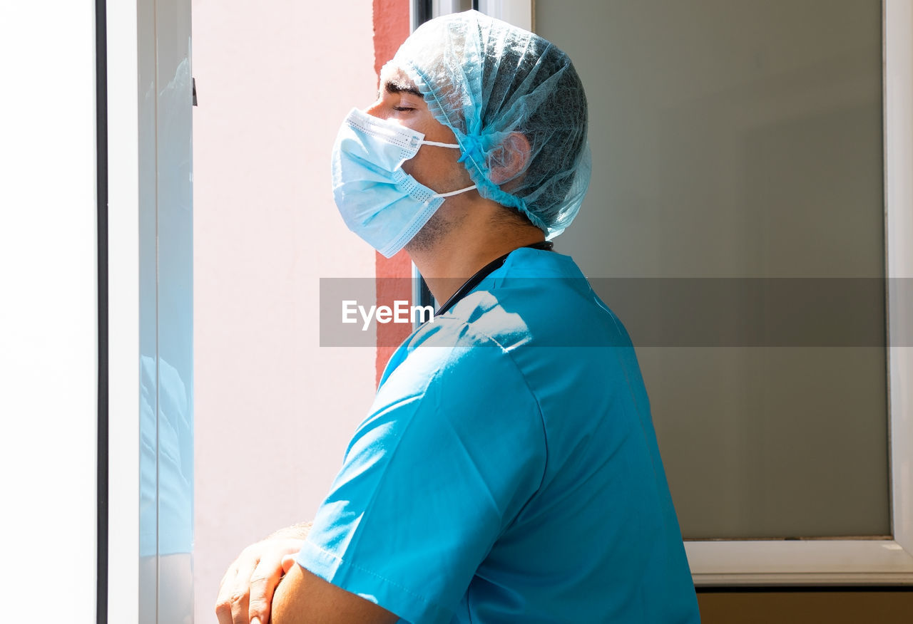 Side view of tired male medic in mask and uniform standing near window in clinic and having break