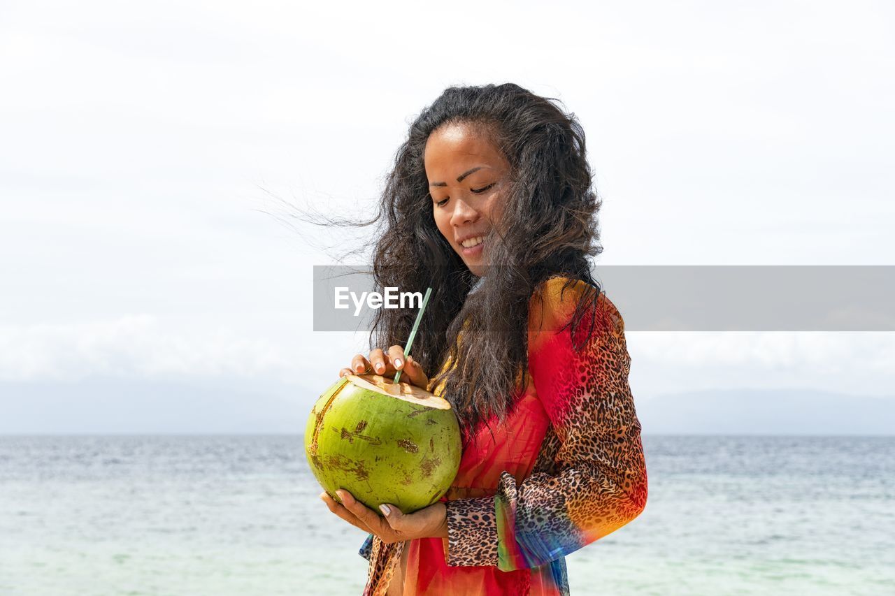 Young woman holding coconut water at beach against sky
