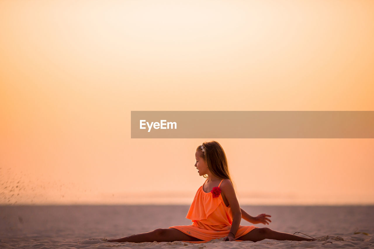 MAN SITTING ON BEACH AGAINST ORANGE SKY