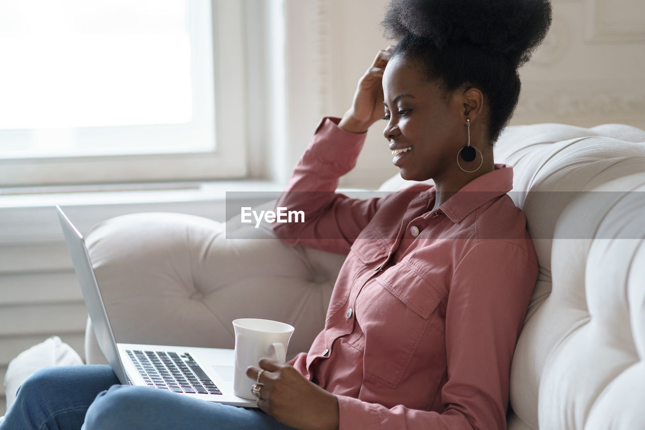 Side view of young woman holding coffee cup while sitting on sofa at home