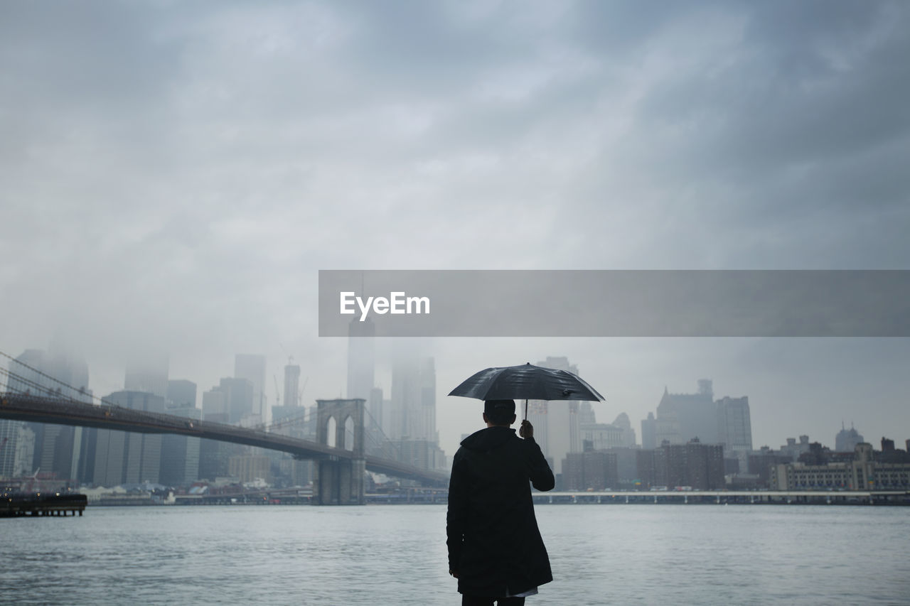 Rear view of man holding umbrella while looking at city view against cloudy sky