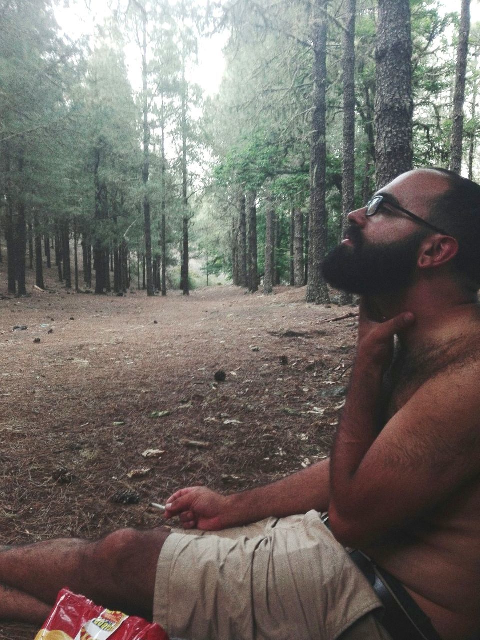 Side view of shirtless man sitting against trees in forest
