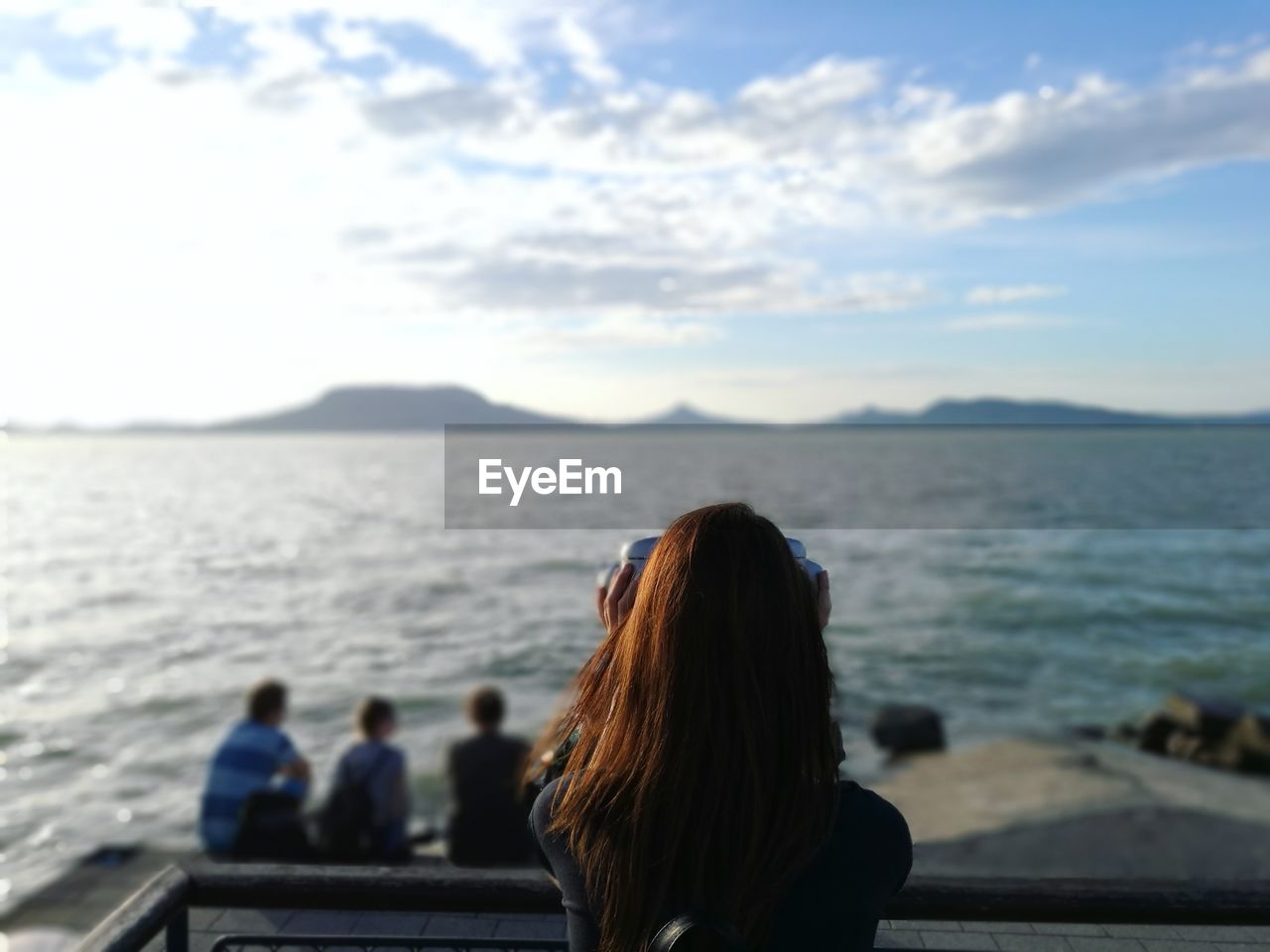 Rear view of young woman looking sea through coin-operated binoculars against sky