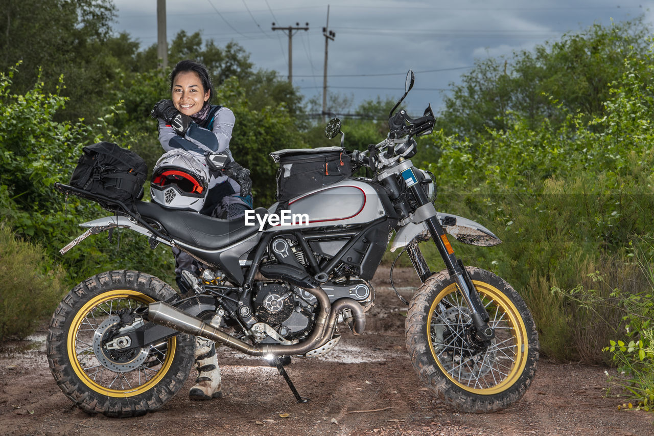 Woman posing behind her scrambler style motorcycle on dirt road