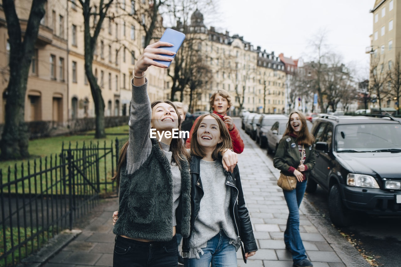 Smiling girl taking selfie through smart phone with friends while standing on sidewalk in city