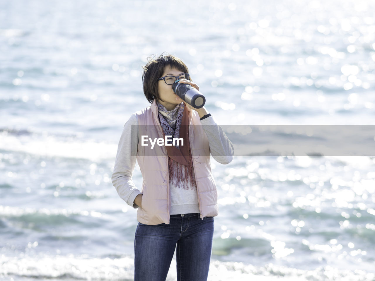 Woman drinks hot tea from metal travel mug. vacation on ocean coast. turquoise water behind woman.