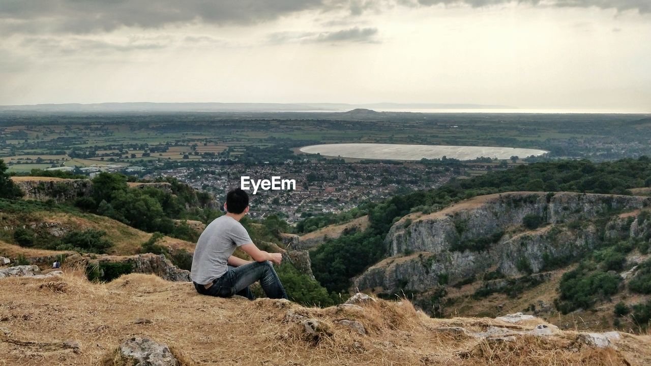 Man sitting on cliff against landscape during sunset