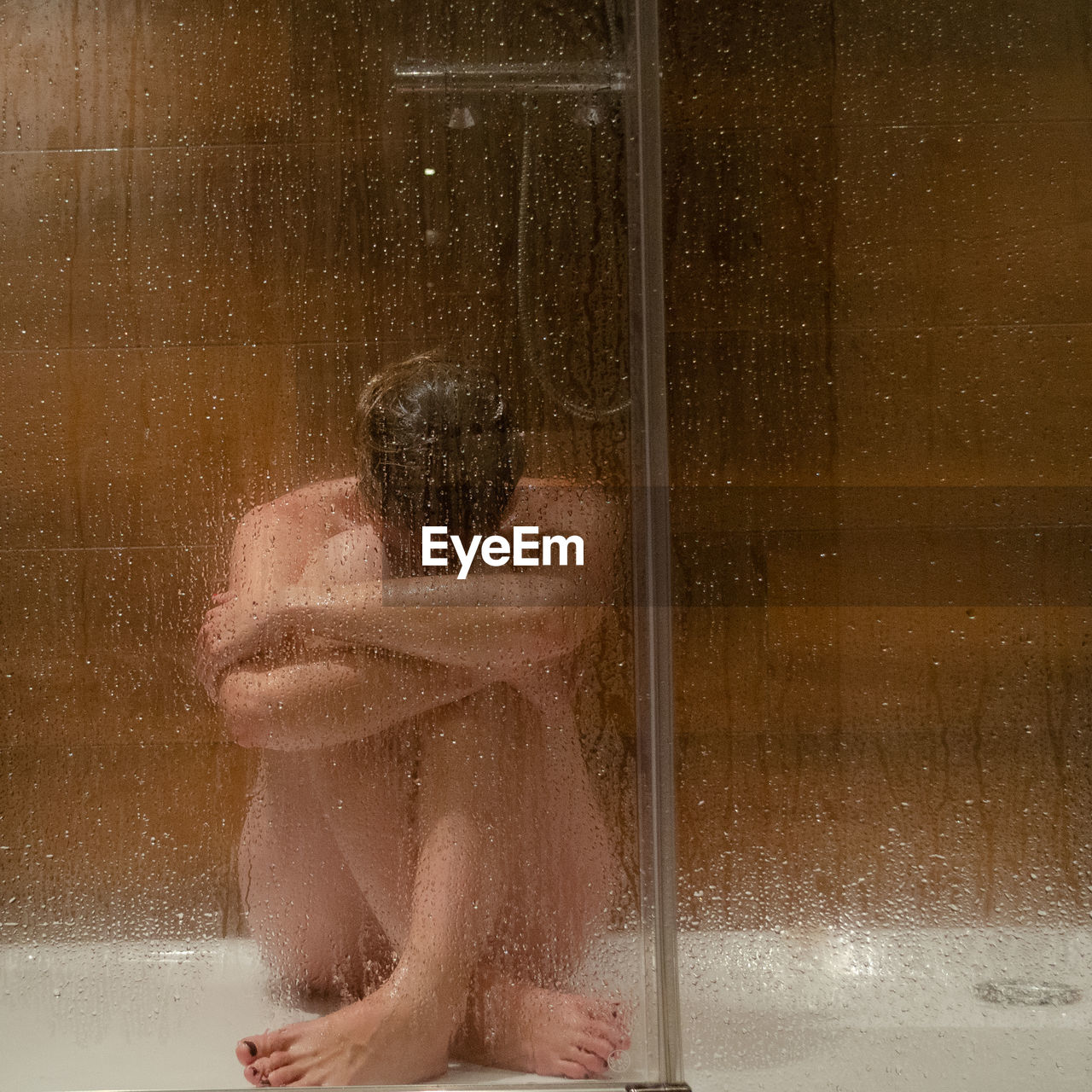 Full length of shirtless depressed woman sitting in bathroom seen through wet glass window