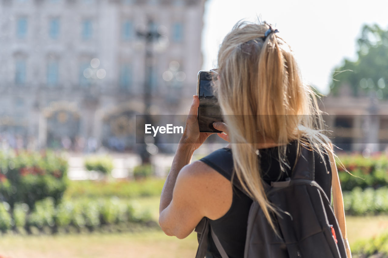 Rear view of a blonde woman taking a smartphone photo of buckingham palace out of focus.