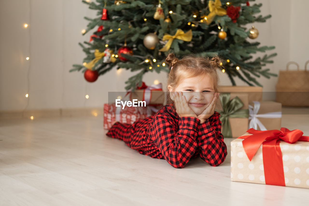 Cute little child girl in red pajama lying on floor near christmas tree with gift boxes at home