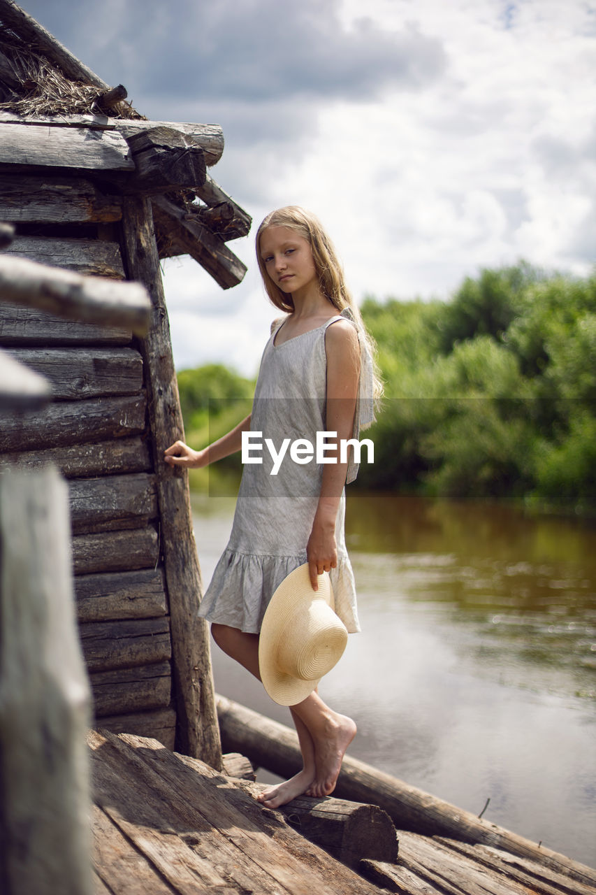A teenage village girl in a dress stands at a wooden house by the river in summer