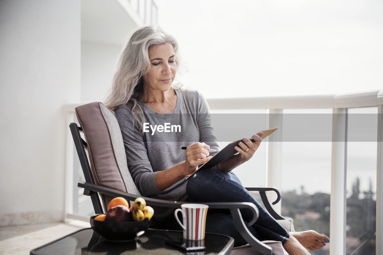 Mature woman writing in book while sitting on chair on balcony