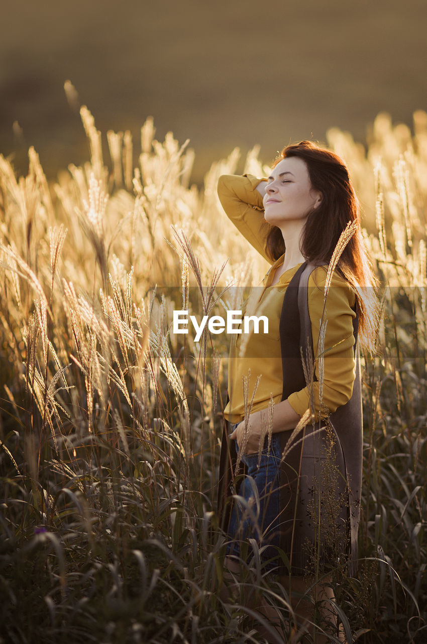 Brunette woman yellow shirt and vest made of eco leather enjoys sunset warm light in field of millet