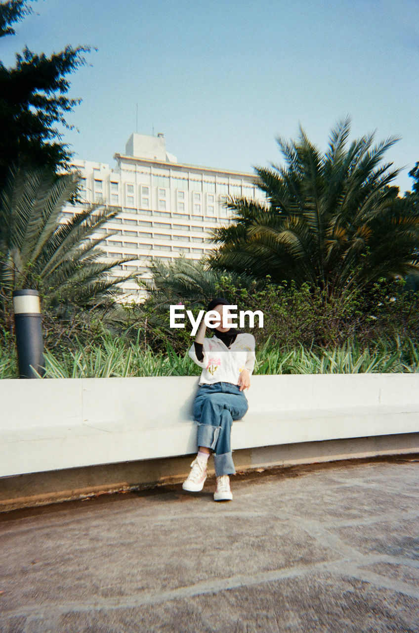 full length, one person, architecture, tree, plant, casual clothing, young adult, vacation, day, adult, footwear, nature, building exterior, built structure, leisure activity, city, sky, palm tree, standing, women, lifestyles, front view, clothing, outdoors, portrait, blue, person, emotion, looking, looking at camera, smiling, happiness