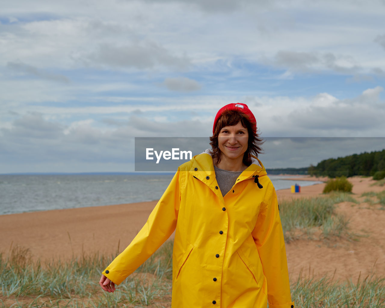 Waist portrait of mature woman in yellow raincoat and red hat walking along beach 