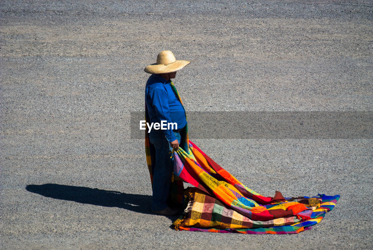 Side view of man wearing sombrero holding multi colored blanket while standing on street