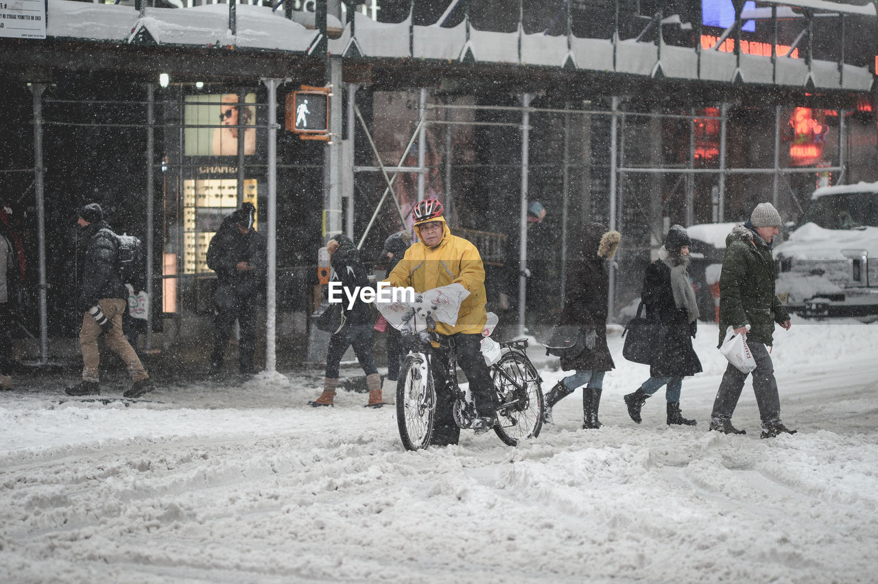 transportation, mid adult, snow, snowing, city, winter, helmet, full length, men, only men, cold temperature, day, real people, building exterior, outdoors, coworker, bicycle, challenge, sport, built structure, motion, adult, people, adults only, road, architecture, corporate business, athlete, teamwork, headwear, one person, warm clothing, young adult