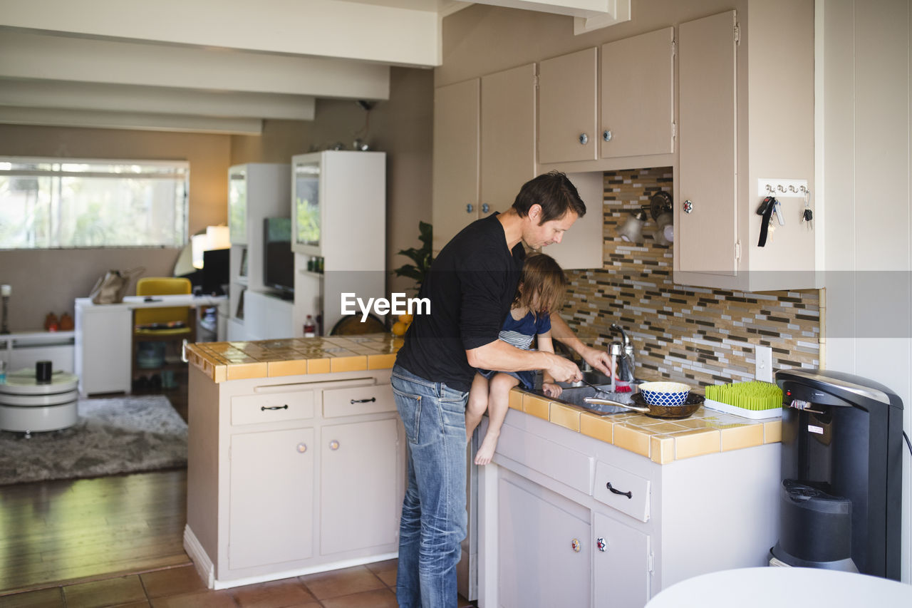 Father washing kitchen utensil while daughter sitting on kitchen counter