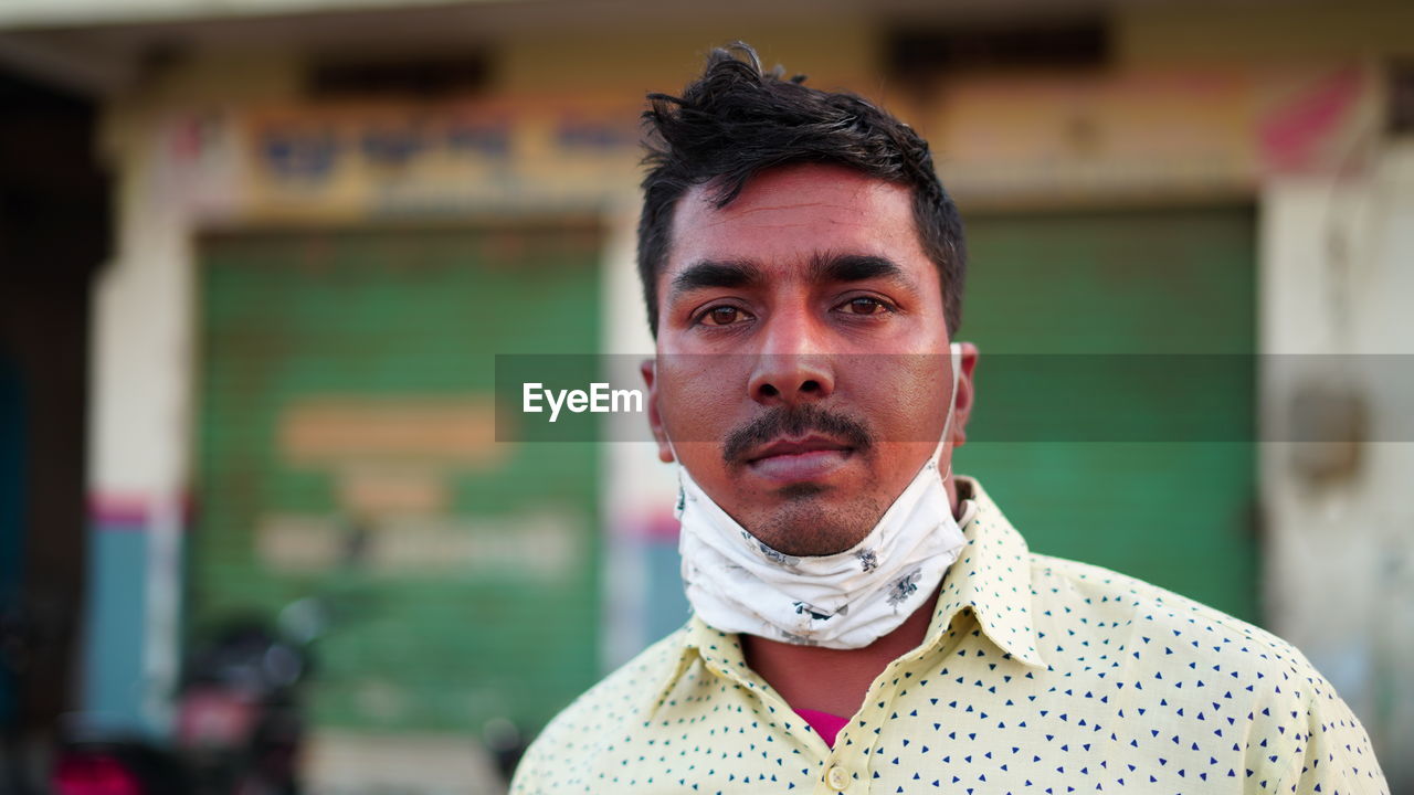 Front shot of indian man wearing mask and shirt over blur market background with serious face.