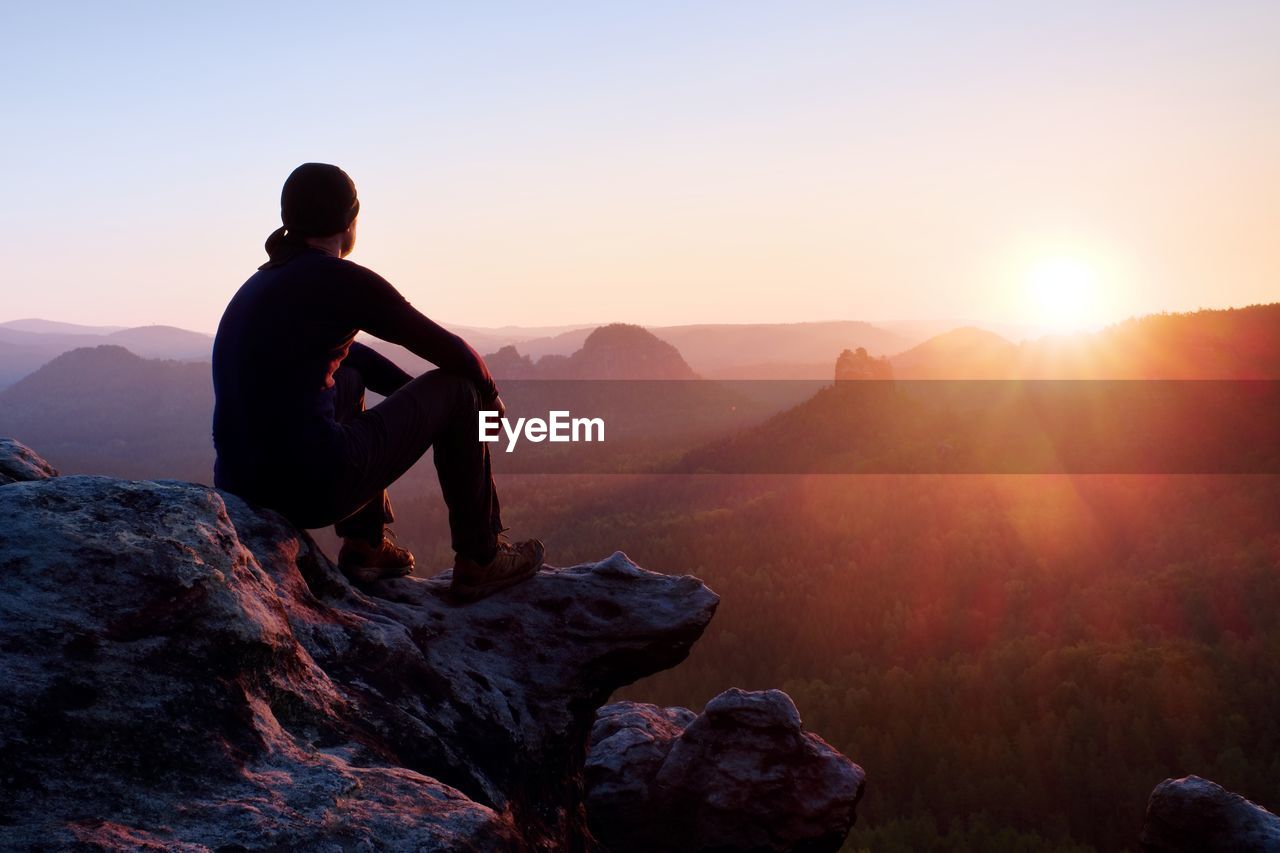 Adult tourist in black trousers, jacket and dark cap sit on cliff's edge and looking to misty hills