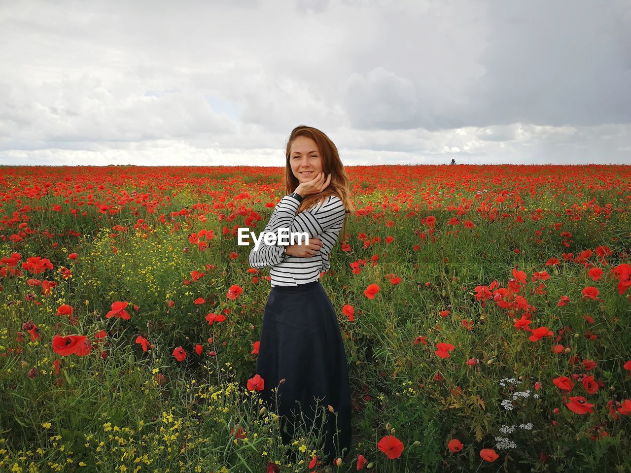 Portrait of woman standing at red poppy farm against cloudy sky