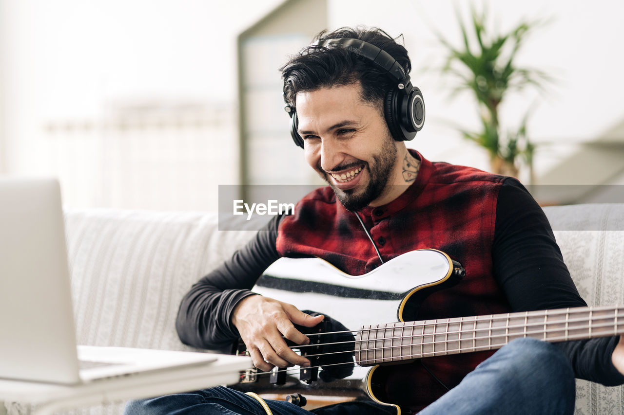 Adult male musician in headphones playing bass guitar against netbook on sofa in living room