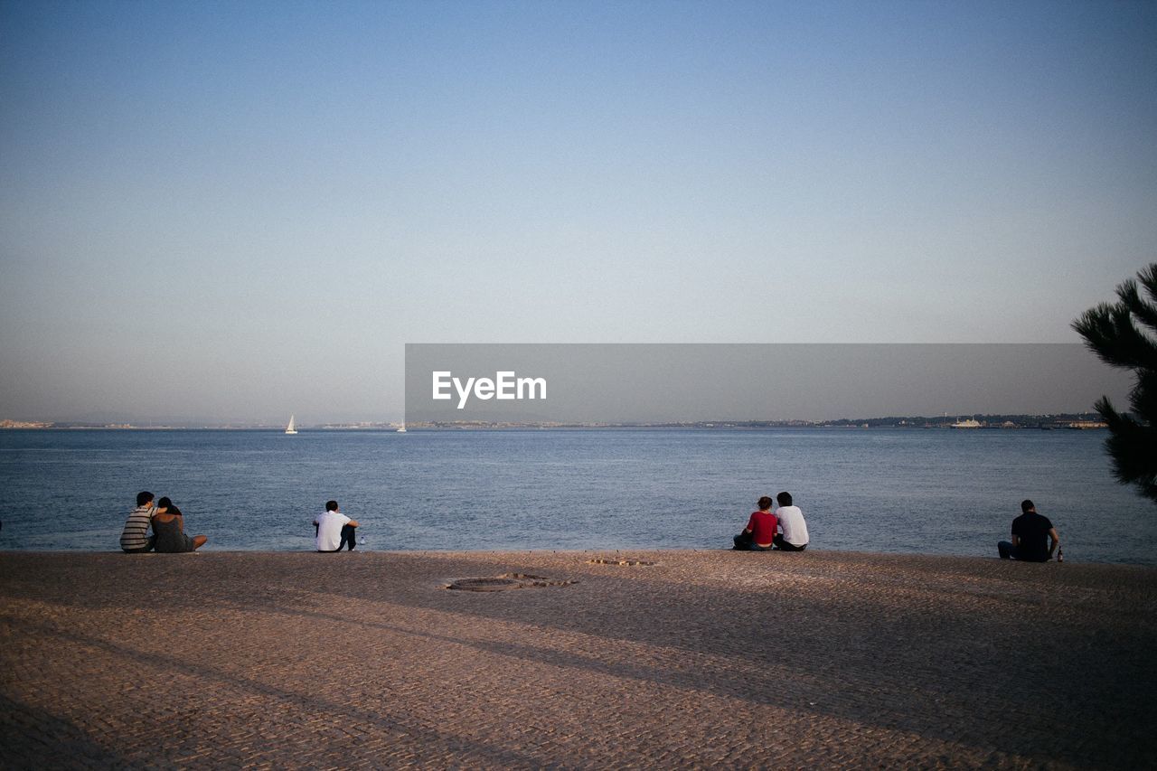 Rear view of people sitting on beach against clear blue sky