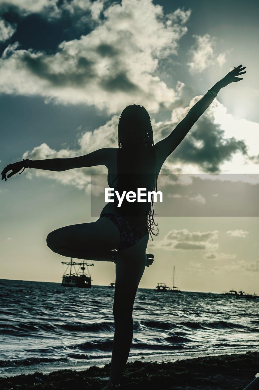 WOMAN WITH ARMS RAISED ON BEACH AGAINST SKY