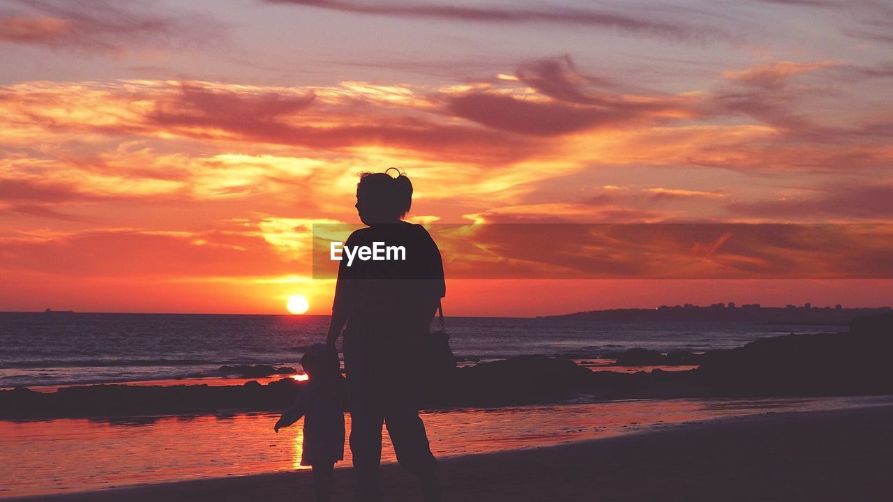 Silhouette mother with daughter standing at beach against sky during sunset