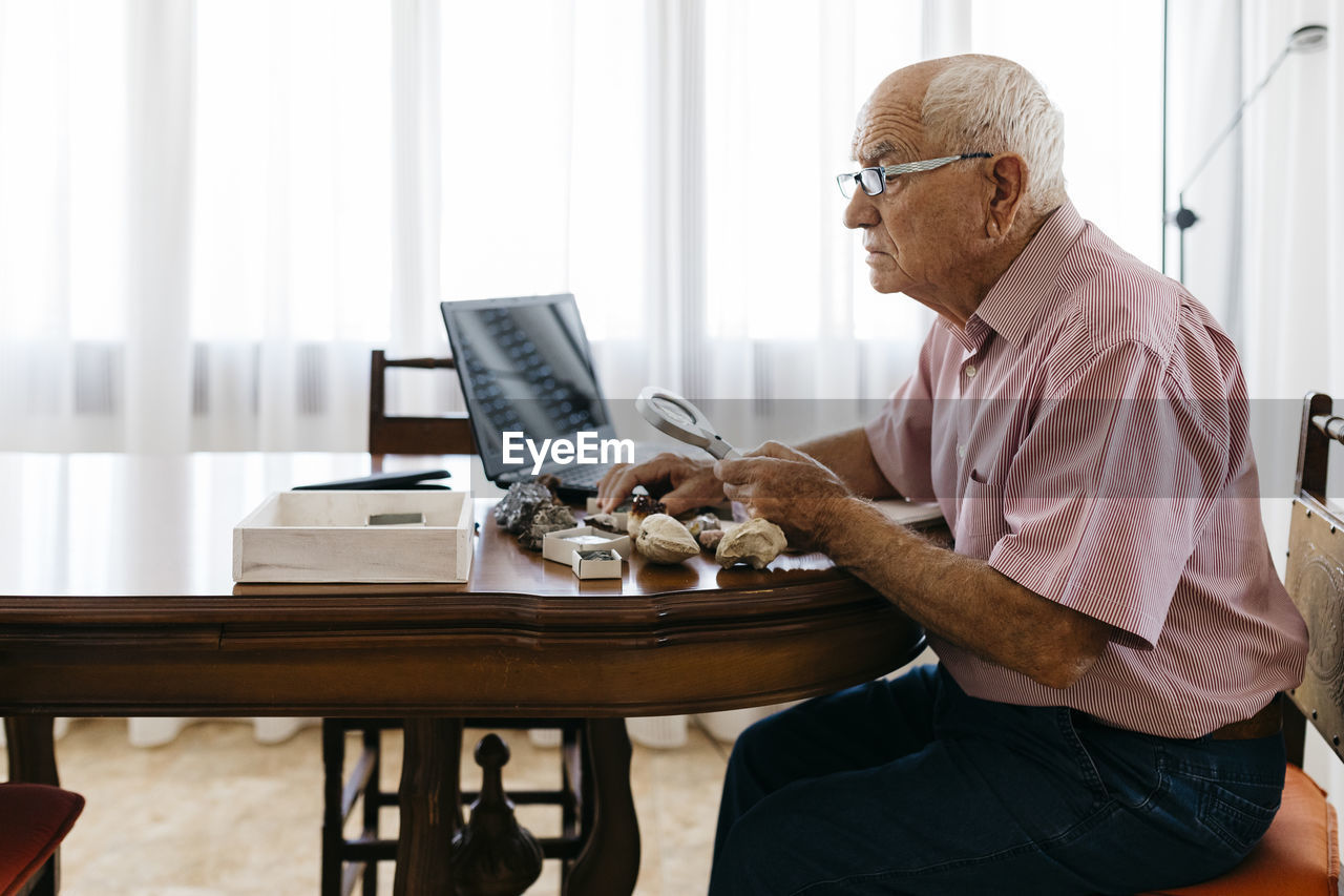 Retired senior man using magnifying glass for research of fossil and mineral at home