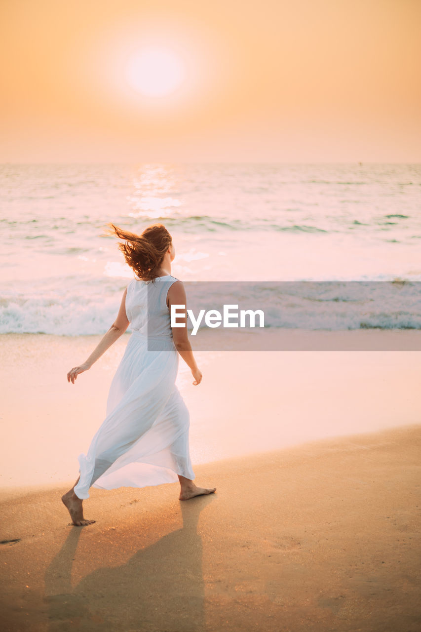 Goa, india. young caucasian woman with fluttering hair in wind in white dress  along seashore sunset 