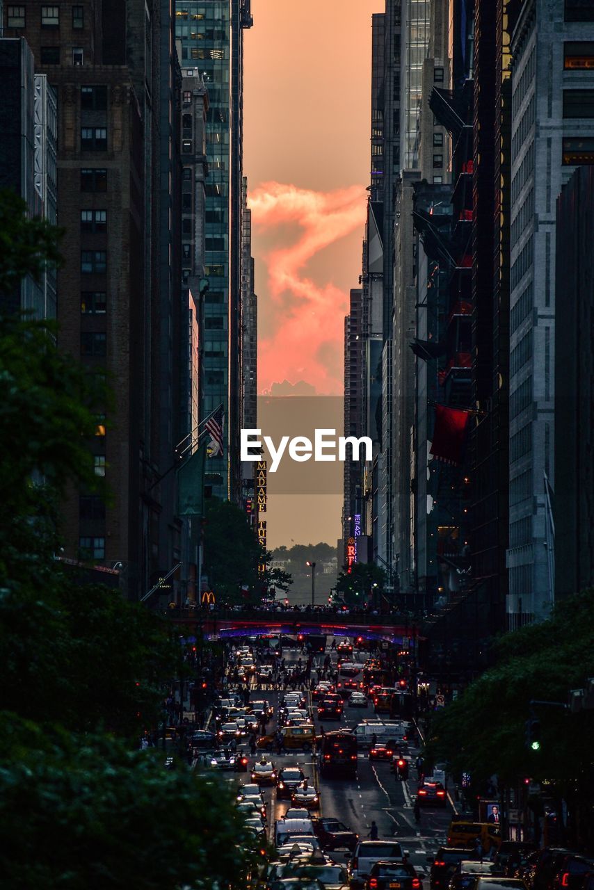 Traffic on city street by buildings against sky during sunset