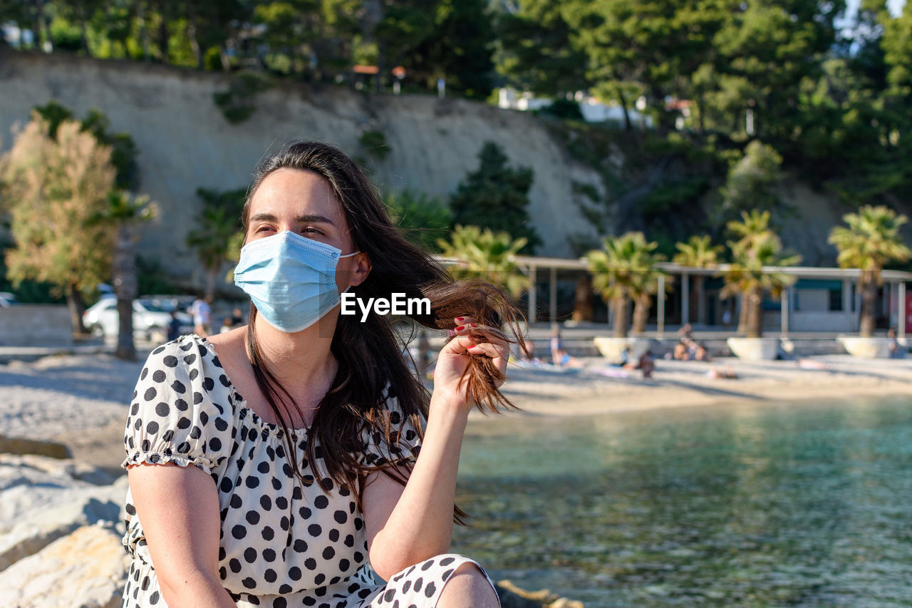 Young woman wearing surgical mask, sitting and relaxing on beach in summer.