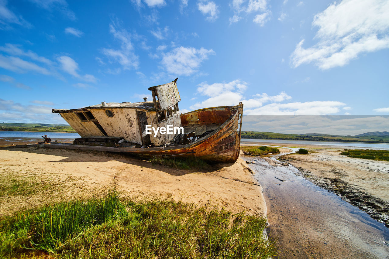 sea, coast, shore, water, sky, nautical vessel, beach, land, transportation, shipwreck, nature, vehicle, cloud, abandoned, boat, mode of transportation, ocean, ship, damaged, watercraft, no people, coastline, environment, sand, scenics - nature, landscape, travel, day, outdoors, bay, rusty, old, rundown, blue, waterway, tranquility, decline, history, deterioration, travel destinations, ruined, non-urban scene, broken, wood, tranquil scene, body of water