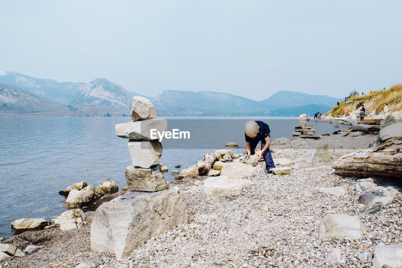 Boy picking up stone while standing on lakeshore against clear sky