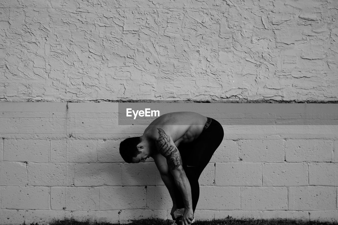 Side view of shirtless man tying shoelace against wall